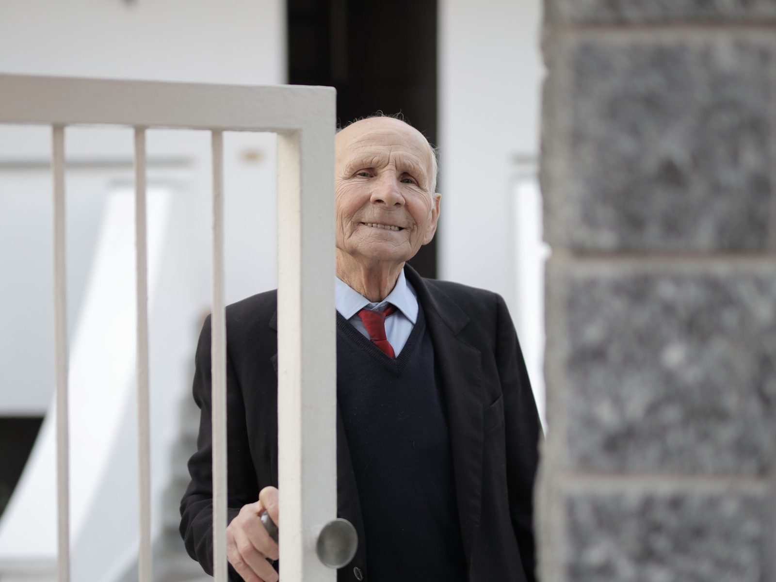 An older man in a suit jacket smiles at his front gate